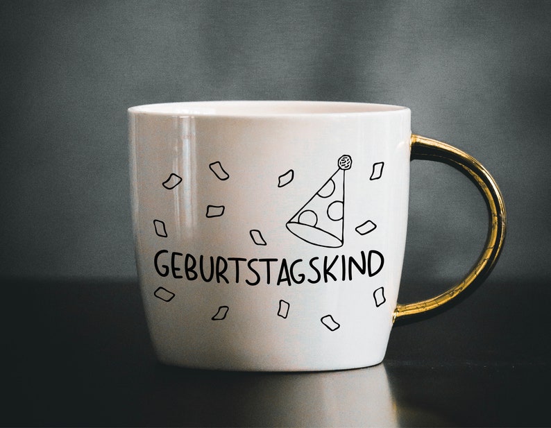May include: White ceramic mug with a gold handle. The mug has a black and white illustration of a party hat and confetti. The text "Geburtstagskind" is printed on the mug.