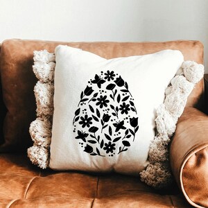 Puede incluir: Una almohada blanca con un dise&ntilde;o floral negro en forma de huevo. La almohada est&aacute; en un sof&aacute; de cuero marr&oacute;n.