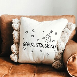 May include: A white throw pillow with a black and white illustration of a party hat and confetti. The text "Geburtstagskind" is printed on the pillow.