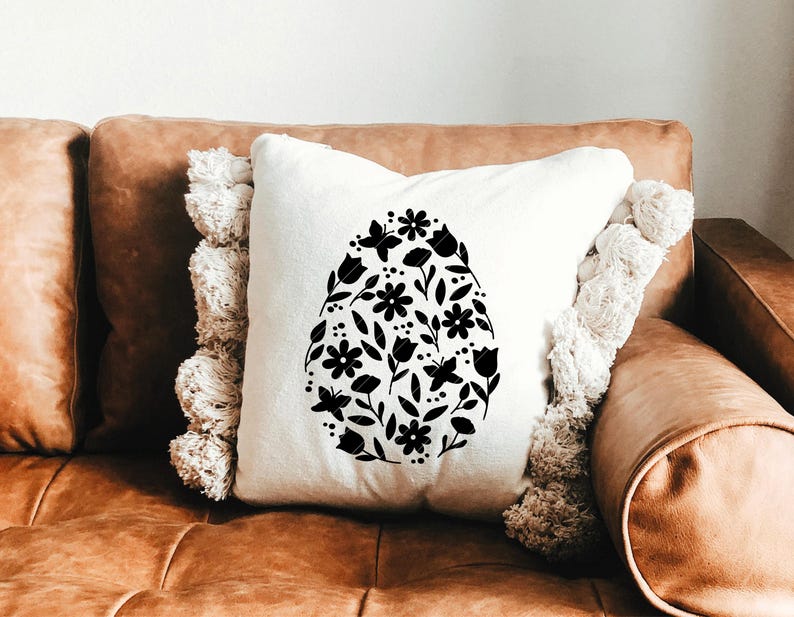 Puede incluir: Una almohada blanca con un dise&ntilde;o floral negro en forma de huevo. La almohada est&aacute; en un sof&aacute; de cuero marr&oacute;n.