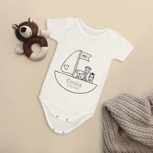 K&ouml;nnte beinhalten: Wei&szlig;er Babybody mit einer schwarz-wei&szlig;en Illustration eines Segelboots mit einem B&auml;ren, einer Giraffe und einem Zebra darauf. Der Text "Taufe" steht auf dem Segel und "Emilia 11.06.2023" steht unter dem Boot.