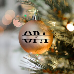 May include: A gold Christmas ornament with the word "OPA" written in black letters with a horizontal line separating the letters. The word "Jan" is written in smaller letters below the line.
