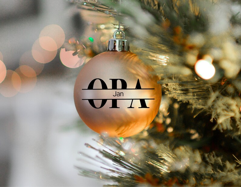 May include: A gold Christmas ornament with the word "OPA" written in black letters with a horizontal line separating the letters. The word "Jan" is written in smaller letters below the line.