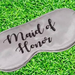 Puede incluir: Una máscara para dormir de satén gris con el texto "Maid of Honor" impreso en negro.