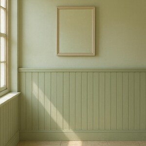 May include: A light green room with a framed mirror hanging on the wall. The lower half of the wall features vertical paneling. A window is visible on the left side, and the floor is tiled. Sunlight streams in from the window.