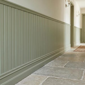 May include: A hallway with sage green wainscoting and a stone tile floor. The walls are painted a light cream color. A door is visible at the end of the hallway, with a small rug in front of it.