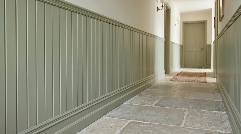May include: A hallway with sage green wainscoting and a stone tile floor. The walls are painted a light cream color. A door is visible at the end of the hallway, with a small rug in front of it.