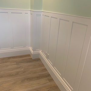 May include: White wainscoting with a raised panel design, featuring a light green wall above and a wood floor below.