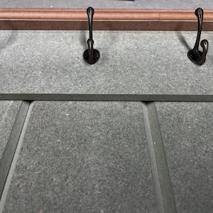 May include: A wall-mounted coat rack with three bronze-colored hooks. The rack has a gray surface with a decorative wooden trim at the top. The hooks are evenly spaced and have a curved design.