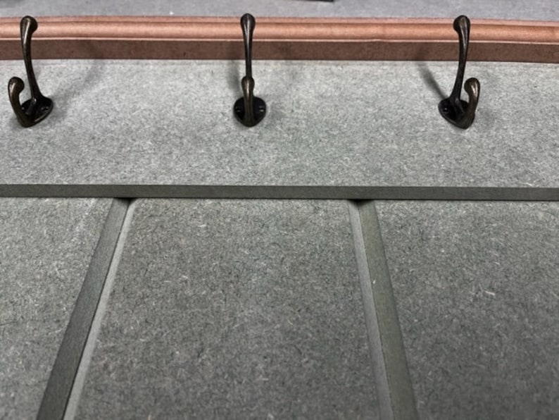 May include: A wall-mounted coat rack with three bronze-colored hooks. The rack has a gray surface with a decorative wooden trim at the top. The hooks are evenly spaced and have a curved design.