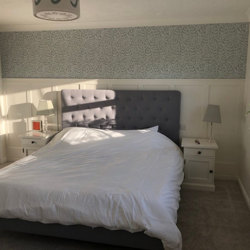 Op de afbeelding: Een wit bed met een grijs hoofdeinde en witte beddengoed. Het hoofdeinde heeft een gestoffeerde vormgeving en het bed staat in een slaapkamer met witte muren en een behang met patroon.