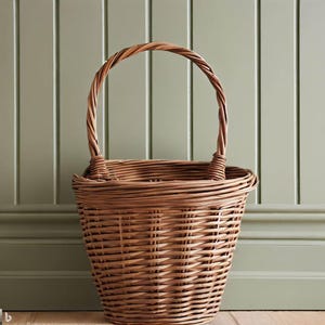 May include: A woven wicker basket with a rounded base and a tall, arched handle. The basket is a warm brown color and has a textured surface. The handle is made of twisted wicker. The basket is empty and sits against a light green wall.