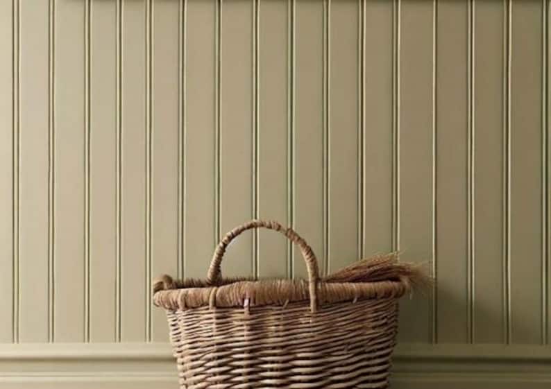 May include: A woven wicker basket with a handle sits in front of a light green vertical panel wall. The basket is filled with dried grasses. The wall and basket create a rustic, farmhouse aesthetic.