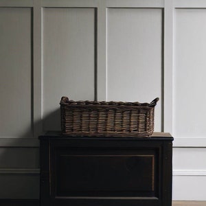 May include: A brown wicker basket with two handles sits on top of a dark brown wooden chest. The chest has a drawer with a dark wood finish. The basket and chest are in front of a white paneled wall.