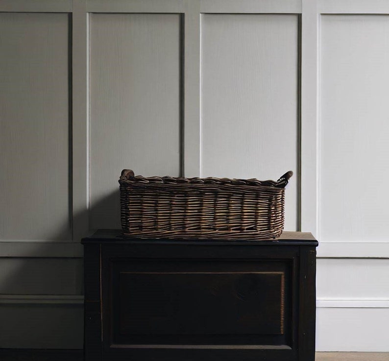 May include: A brown wicker basket with two handles sits on top of a dark brown wooden chest. The chest has a drawer with a dark wood finish. The basket and chest are in front of a white paneled wall.