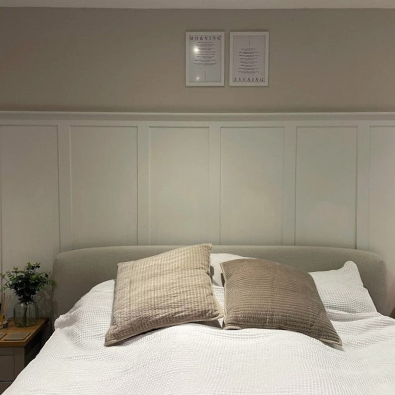 Op de afbeelding: Een wit bed met een wit hoofdbord en een wit dekbedovertrek. Twee bruine kussens liggen op het bed. Het hoofdbord heeft een wit lambriseringontwerp. Twee ingelijste prints hangen aan de muur boven het hoofdbord. De print aan de linkerkant zegt "Ochtend" en de print aan de rechterkant zegt "Avond".