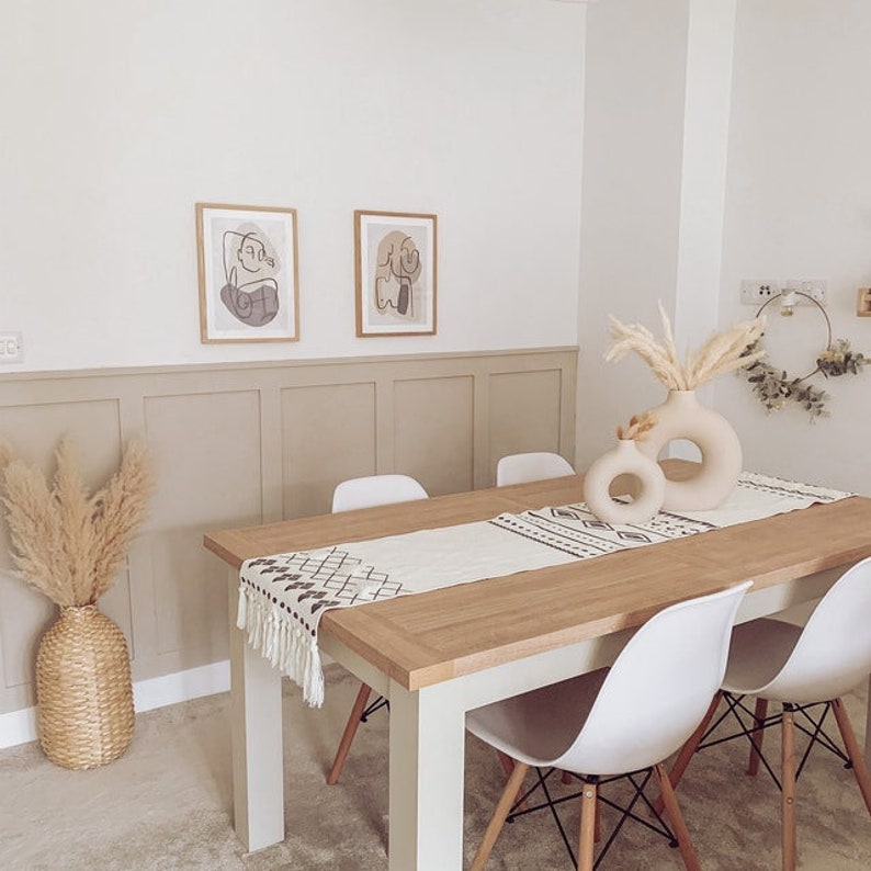 May include: A light wood dining table with a white runner and a geometric pattern. The table is set with four white chairs and a vase with dried pampas grass. Two abstract line drawings are on the wall above the table.