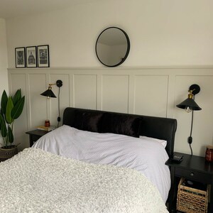 May include: A white bed with a fluffy white blanket and black pillows. The bed is in a bedroom with white walls and a black and gold wall sconce on each side of the bed. There is a plant in a brown pot on the floor next to the bed.