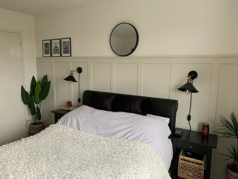 May include: A white bed with a fluffy white blanket and black pillows. The bed is in a bedroom with white walls and a black and gold wall sconce on each side of the bed. There is a plant in a brown pot on the floor next to the bed.