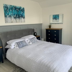 May include: A white bed with a gray headboard and white bedding. There are two pillows with geometric patterns on the bed. The bed is in a bedroom with a dresser and a mirror on the wall.