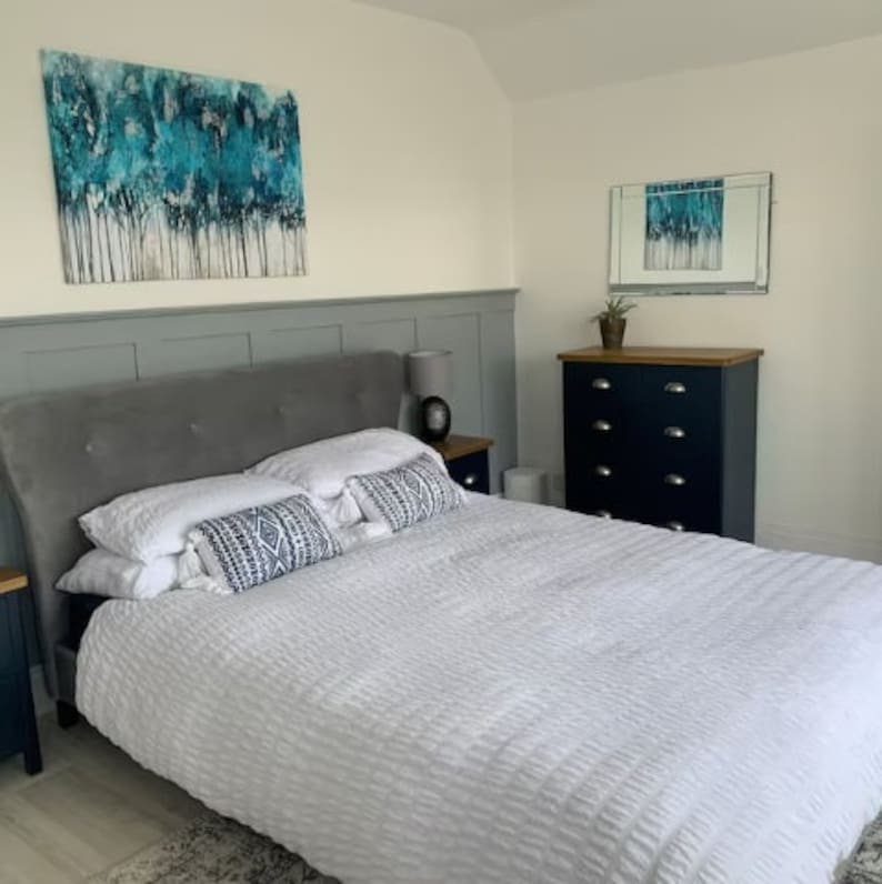May include: A white bed with a gray headboard and white bedding. There are two pillows with geometric patterns on the bed. The bed is in a bedroom with a dresser and a mirror on the wall.