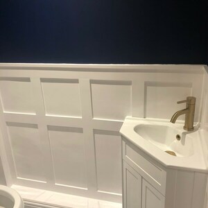 May include: A white bathroom vanity with a corner sink and gold faucet. The vanity has two doors and is set against a white wainscot wall with a dark blue accent wall above. The floor is tiled with a white and blue patterned tile.