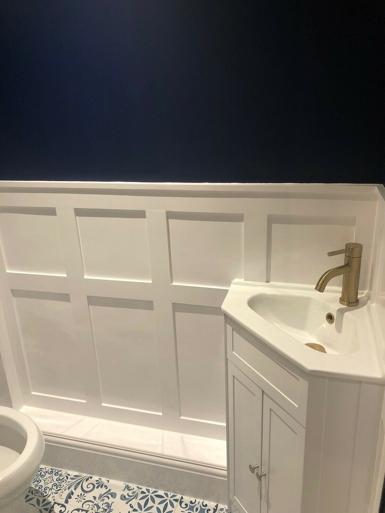 May include: A white bathroom vanity with a corner sink and gold faucet. The vanity has two doors and is set against a white wainscot wall with a dark blue accent wall above. The floor is tiled with a white and blue patterned tile.