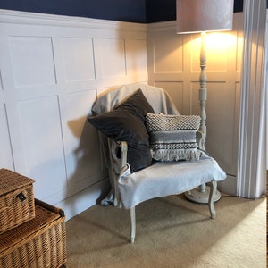 May include: A white armchair with a gray throw blanket and two pillows, one black and one black and white patterned, sits in a corner of a room with white wainscoting and a floor lamp with a white lampshade.