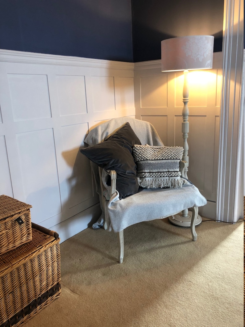 May include: A white armchair with a gray throw blanket and two pillows, one black and one black and white patterned, sits in a corner of a room with white wainscoting and a floor lamp with a white lampshade.
