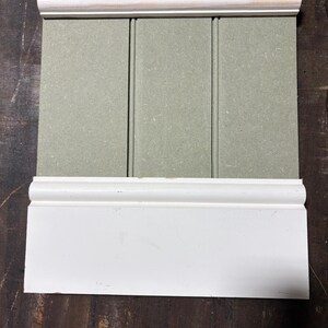 May include: A section of a wall paneling system, featuring a light green vertical panel with white trim at the top and bottom. The top trim includes a natural wood edge. The paneling is designed for interior use.