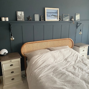May include: A bedroom with a dark blue wall and white wood paneling. A wicker headboard bed with white bedding is centered in the room. Two white nightstands with drawers are on either side of the bed.