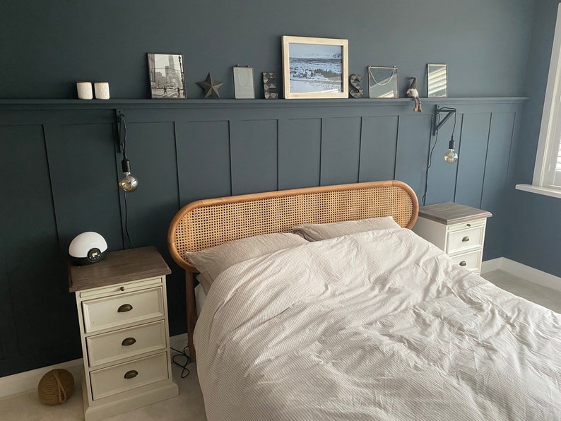 May include: A bedroom with a dark blue wall and white wood paneling. A wicker headboard bed with white bedding is centered in the room. Two white nightstands with drawers are on either side of the bed.