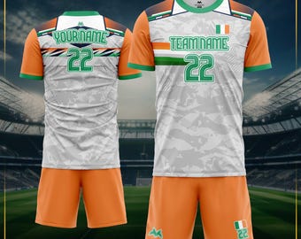 May include: A soccer uniform set featuring a white and gray jersey with orange sleeves and shorts. The jersey has the text "YOURNAME" and "TEAMNAME" with the number "22" in green. The shorts are orange with a green logo and the number "22".