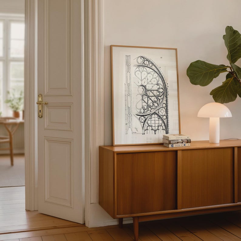 Puede incluir: Un mueble de madera con puertas correderas exhibe un dibujo arquitect&oacute;nico enmarcado y una l&aacute;mpara blanca con forma de hongo. La obra de arte es un plano en blanco y negro de una ventana de catedral. Una planta verde est&aacute; en el fondo.