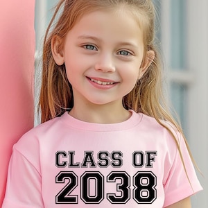May include: A young girl wearing a pale pink t-shirt with the text "CLASS OF 2038" in black block letters. The girl is smiling and leaning against a pink wall. The shirt is short-sleeved.