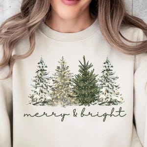 May include: Cream-coloured sweatshirt with a watercolour design of fir trees in green and white. The words "merry & bright" are written in a delicate script below the trees. The sweatshirt is a neutral colour.