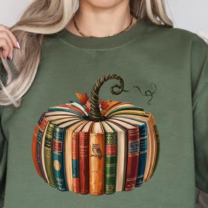 May include: An olive green sweatshirt with a pumpkin design made of books. The pumpkin has a curly stem and autumn leaves. The books are various colours, including orange, red, and green. An owl is visible on one of the book spines.