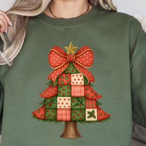 May include: An olive green sweatshirt with a festive Christmas tree design. The tree is made of red and green patchwork squares, a red bow, and a gold star. The trunk is brown. The sweatshirt is a relaxed, casual style, perfect for the holiday season.