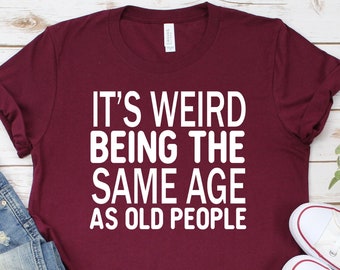 Könnte beinhalten: Ein maroonfarbenes T-Shirt mit weißem Text, der "Es ist komisch, das gleiche Alter wie alte Leute zu haben" lautet. Das Shirt liegt flach auf einer weißen Holzoberfläche mit einer blauen Jeans und weißen Turnschuhen.