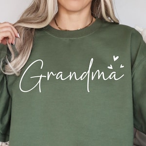 May include: An olive green sweatshirt featuring the word "Grandma" in white script, with two small white hearts. The sweatshirt has a crew neck design.