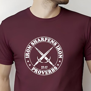 Peut inclure: T-shirt bordeaux avec un motif circulaire blanc. Le motif présente des épées croisées et le texte "IRON SHARPENS IRON 27:17 PROVERBES". Le t-shirt a des manches courtes et un col rond.