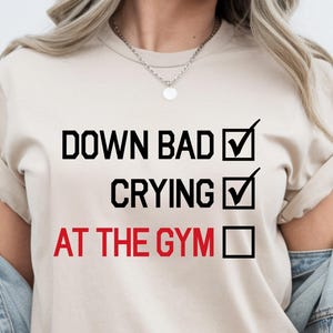 Peut inclure: Un t-shirt beige à manches courtes avec le texte noir DOWN BAD accompagné d'une coche, CRYING avec une coche, et le texte rouge AT THE GYM à côté d'une case vide. Le t-shirt est porté avec une veste en jean bleu clair.