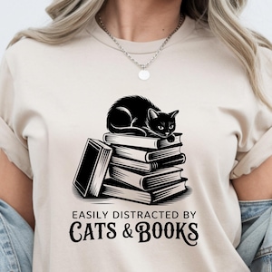 May include: A beige short-sleeved t-shirt featuring a black graphic of a cat resting on a stack of books. Below the illustration, the text reads EASILY DISTRACTED BY CATS & BOOKS in a bold, stylised font. The shirt is worn with a light blue denim jacket.