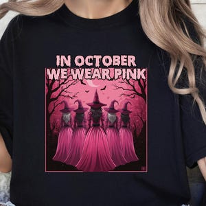 Peut inclure: T-shirt noir avec l'inscription "IN OCTOBER WE WEAR PINK" au-dessus d'une illustration de cinq sorcières en robes et chapeaux roses. Les sorcières se tiennent devant un arbre rose et une pleine lune.