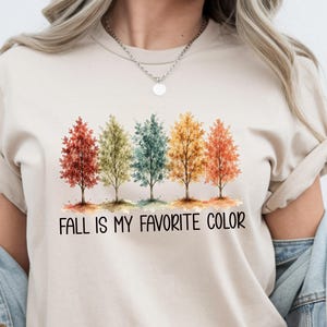 May include: A beige short-sleeved t-shirt featuring a graphic of five trees in varying autumn colours including red, green, teal, yellow, and orange. Below the trees, the text reads "FALL IS MY FAVORITE COLOR" in a simple black sans-serif font.