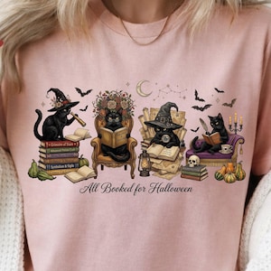 May include: A light pink t-shirt featuring a graphic of four black cats in witch hats reading books, surrounded by pumpkins, bats, and a crescent moon. The text "All Booked for Halloween" is printed in black script below the illustration.