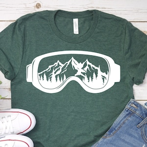 May include: A green t-shirt featuring a white graphic of ski goggles with mountains and trees inside the lenses. The t-shirt is lying on a white wooden surface with a pair of white trainers and blue denim shorts.