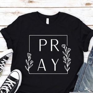 May include: Black t-shirt with white text that says "PRAY" inside a square with floral accents.