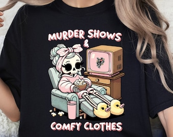 Könnte beinhalten: Schwarzes T-Shirt mit einem Grafikdruck eines Skeletts, das einen rosa Schlafanzug trägt, fernsieht und Popcorn isst. Der Text auf dem Shirt lautet "Murder Shows & Comfy Clothes".