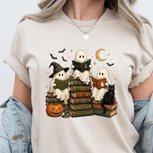 May include: A beige t-shirt featuring a graphic print of three ghosts sitting on a stack of books. The ghosts are reading, accompanied by a black cat, a jack-o'-lantern, a candle, bats, and a crescent moon. The design uses orange, green, and black.